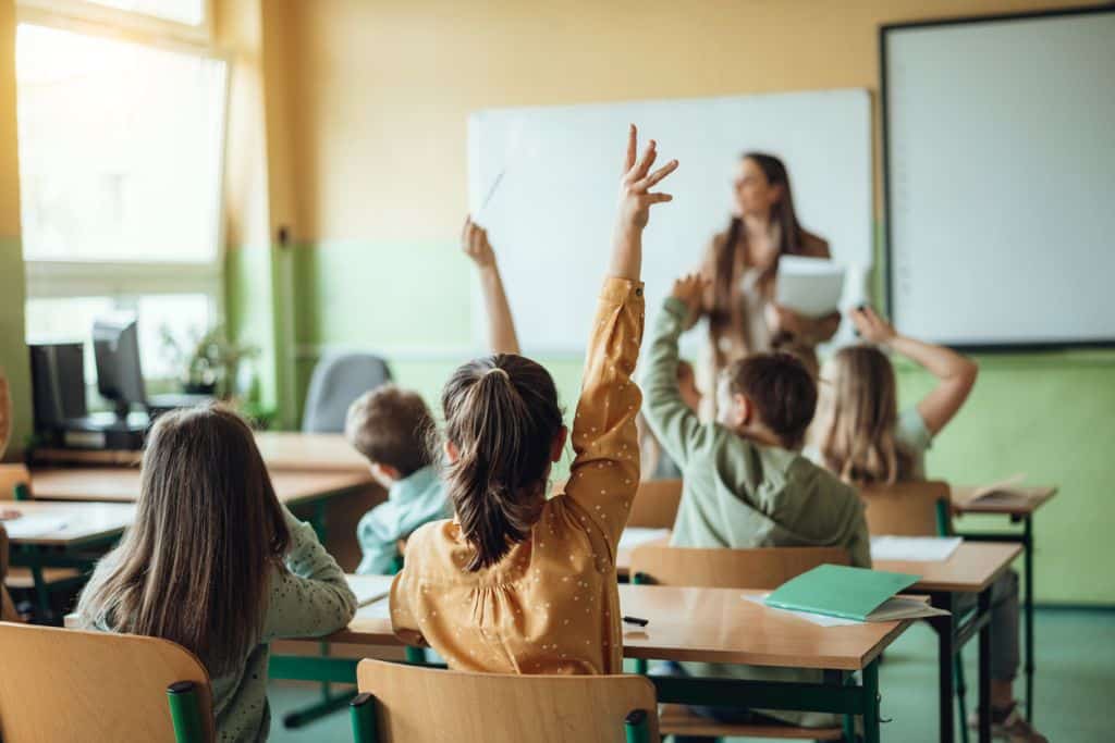 Students seated at desks raise their hands in a classroom while a teacher stands near a whiteboard and projector screen.