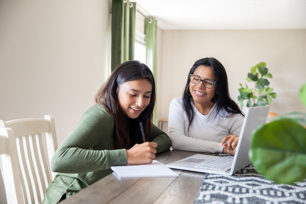 A woman in a green sweater writes in a notebook beside another woman using a laptop at a dining table, with a chair, curtains, and houseplants in the background.