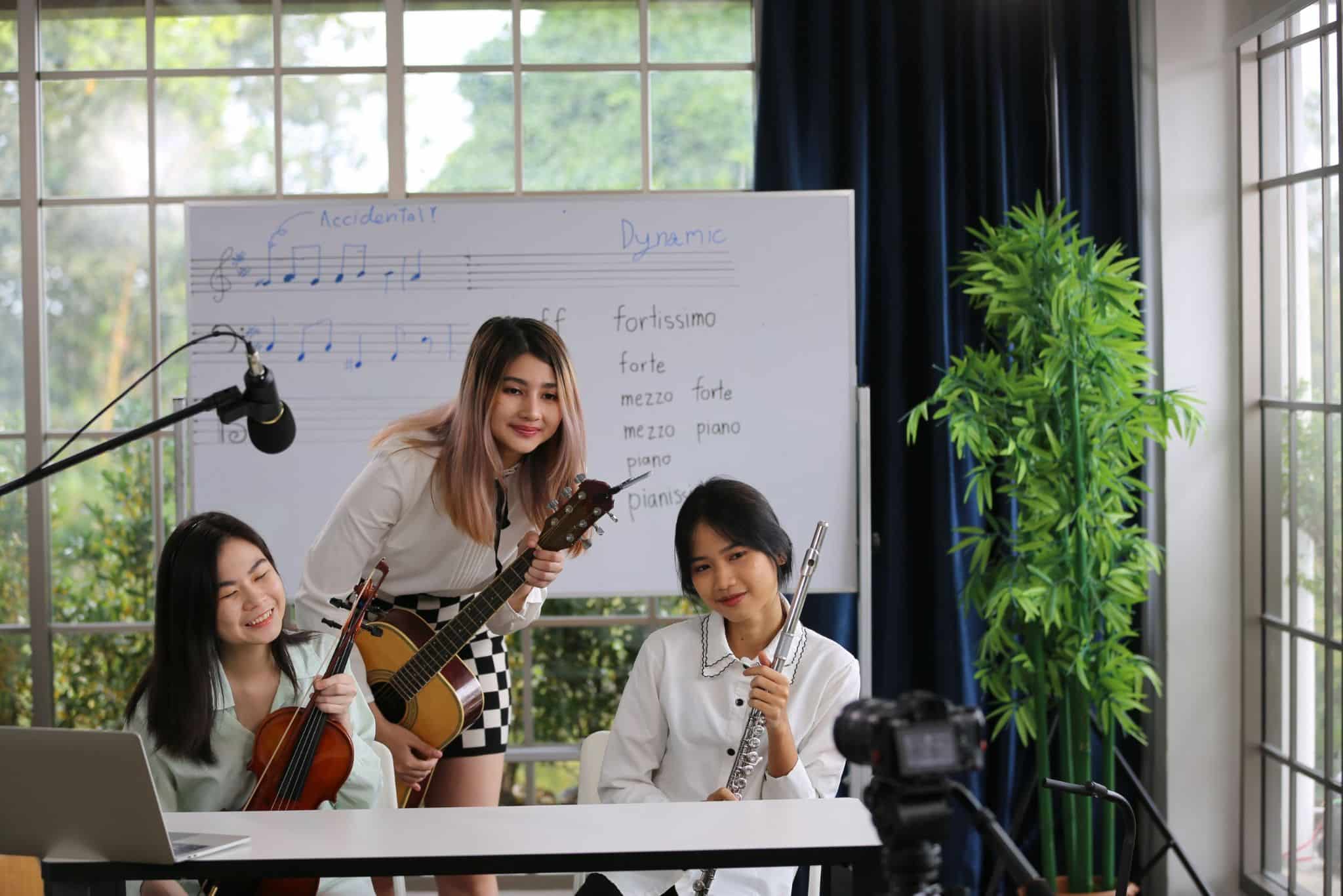 Using Video to Strengthen Family & Community Engagement 1 - MyVideoSpot Three women in a music studio pose with instruments, including a violin and a flute, in front of a whiteboard displaying musical notes and terms. The setting is well-lit with green plants and equipment indicative of an educational video platform for MyVideoSpot Updates.
