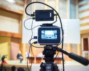 A video camera set up on a tripod, displaying a view of a stage in an auditorium. The camera is connected to various cables, capturing a discussion or presentation, relevant for the MyVideoSpot year in review and MyVideoSpot Updates on the educational video platform.