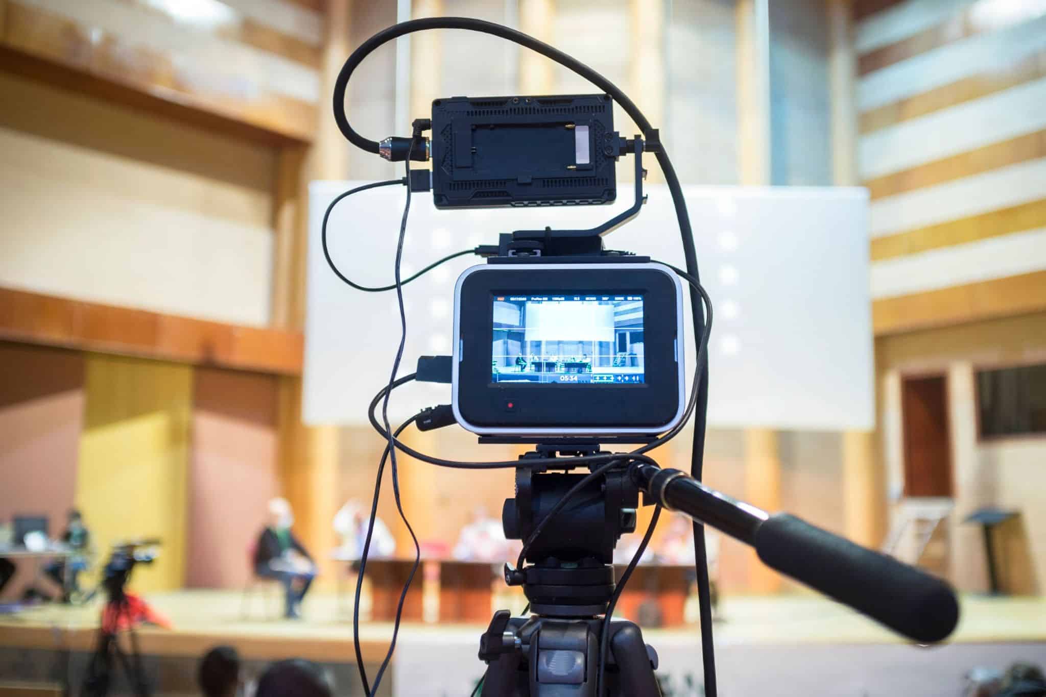 A video camera set up on a tripod, displaying a view of a stage in an auditorium. The camera is connected to various cables, capturing a discussion or presentation, relevant for the MyVideoSpot year in review and MyVideoSpot Updates on the educational video platform.