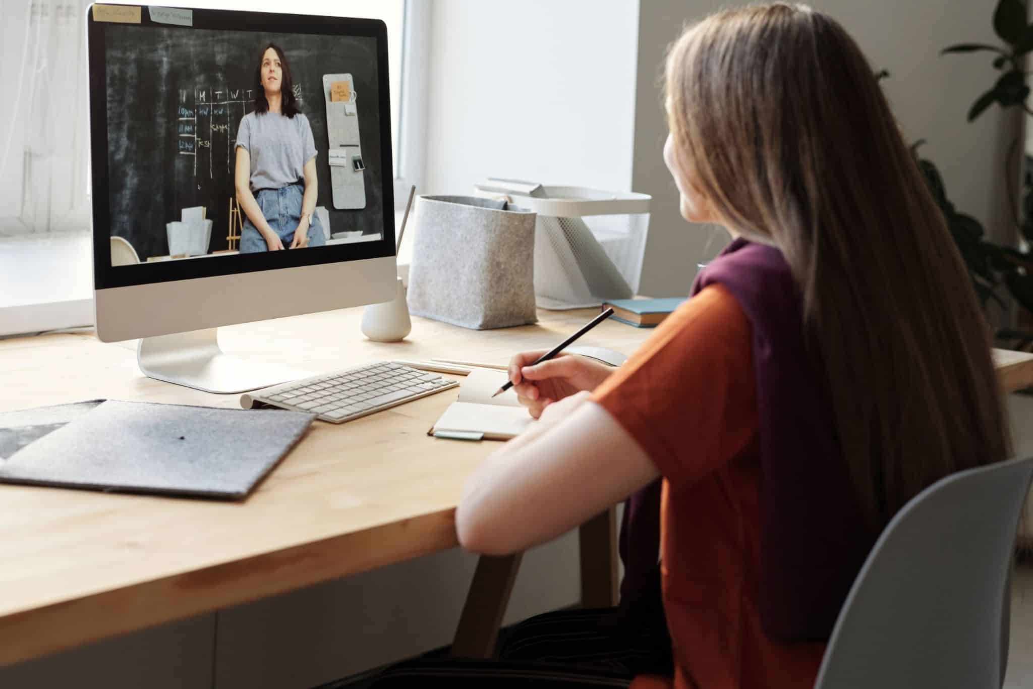 Why Schools Need a Unified Video Platform 1 - MyVideoSpot A woman seated at a desk, taking notes while watching a video on a computer screen. The video shows another woman talking, with a classroom background. The workspace includes a few notebooks and a pencil, indicating use of the educational video platform MyVideoSpot Updates.