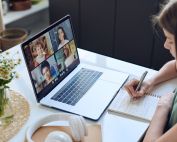A woman seated at a table is taking notes while participating in a video call on her laptop. The screen displays six participants in a grid format. Nearby, a vase of flowers and a pair of headphones are visible. This illustrates MyVideoSpot Updates, an educational video platform.