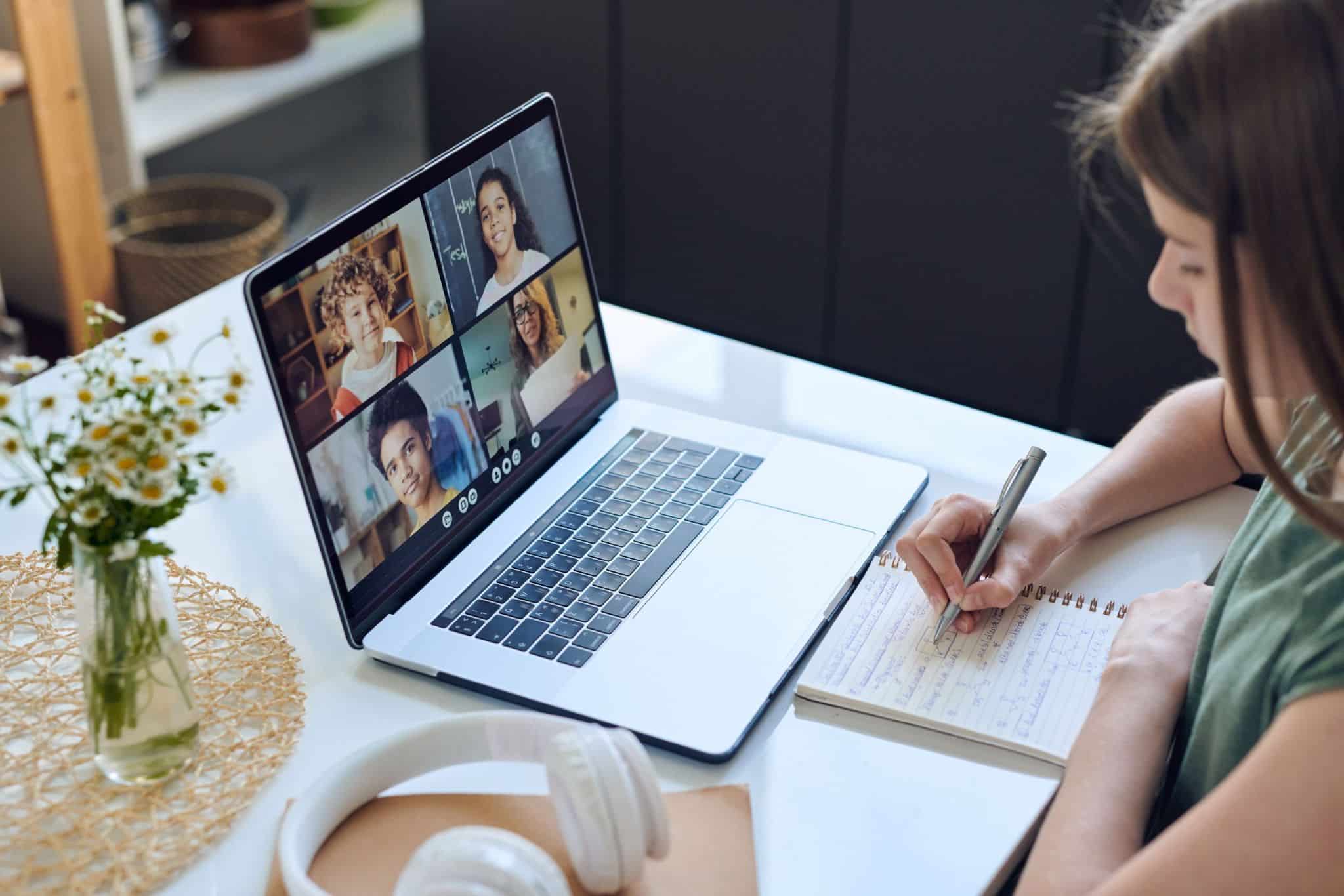 A woman seated at a table is taking notes while participating in a video call on her laptop. The screen displays six participants in a grid format. Nearby, a vase of flowers and a pair of headphones are visible. This illustrates MyVideoSpot Updates, an educational video platform.
